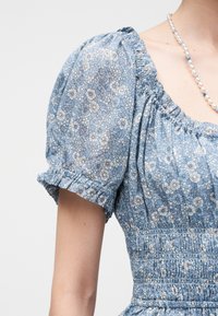 Μπλε floral κοντό φόρεμα με φουσκωτά μανίκια, με συσσωρευμένες λεπτομέρειες και περιδέραιο με πέρλες σε ανοιχτό φόντο.