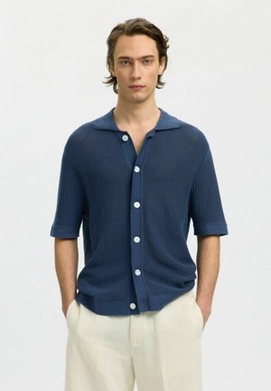 Vállig érő hajú fiatal férfi kék, gombos kötött rövid ujjú inget és világos bézs nadrágot visel, kezét zsebében áll.