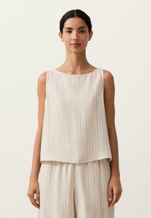 Žena s tmavými vlasmi v bezrukávovom svetlo béžovom topánke s tenkými pruhmi a zodpovedajúcich voľných nohaviciach na jednoduchom béžovom pozadí.