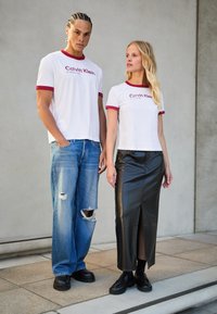 Dvi modeliai vilki atitinkančius baltus marškinėlius su raudonu apvadu, ant kurių užrašas "Calvin Klein". Viena jų derina marškinėlius su suplyšusiais mėlyniais džinsais, o kita - su juoda odine sijoną.