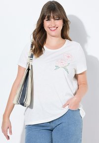 Camiseta blanca de manga corta, con bordados florales en rosa en el lado izquierdo del pecho. Combinada con unos jeans de mezclilla azul y un bolso a rayas beige.