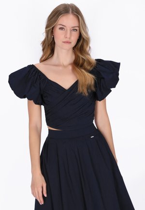 Młoda kobieta z rozpuszczonym falującym włosami nosi czarną sukienkę z dużymi bufkami i dopasowaną, plisowaną spódnicą na białym tle.