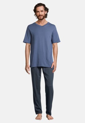 Man staat blootsvoets, draagt een blauw T-shirt met korte mouwen en V-hals en donkere pantalon met fijne strepen tegen een effen lichte achtergrond.