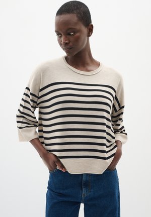 Pull en beige et noir à rayures avec épaules tombantes, col rond et poignets côtelés. Associé à un jean bleu. Texture lisse.