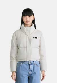 Giubbotto trapuntato bianco con colletto alto, dotato di una patch logo nera, tasche laterali e design corto. La texture presenta un motivo geometrico.