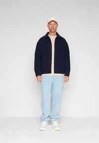 Marine-blå jakke med knapper og to forlommer, båret over en beige skjorte og parret med lysblå jeans og hvide sneakers. Beige kasket på hovedet.