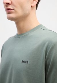 Zelená bavlněná mikina s kulatým výstřihem, krátkými rukávy a lesklým černým logem "BOSS" na hrudi. Hladká textura, volný střih.