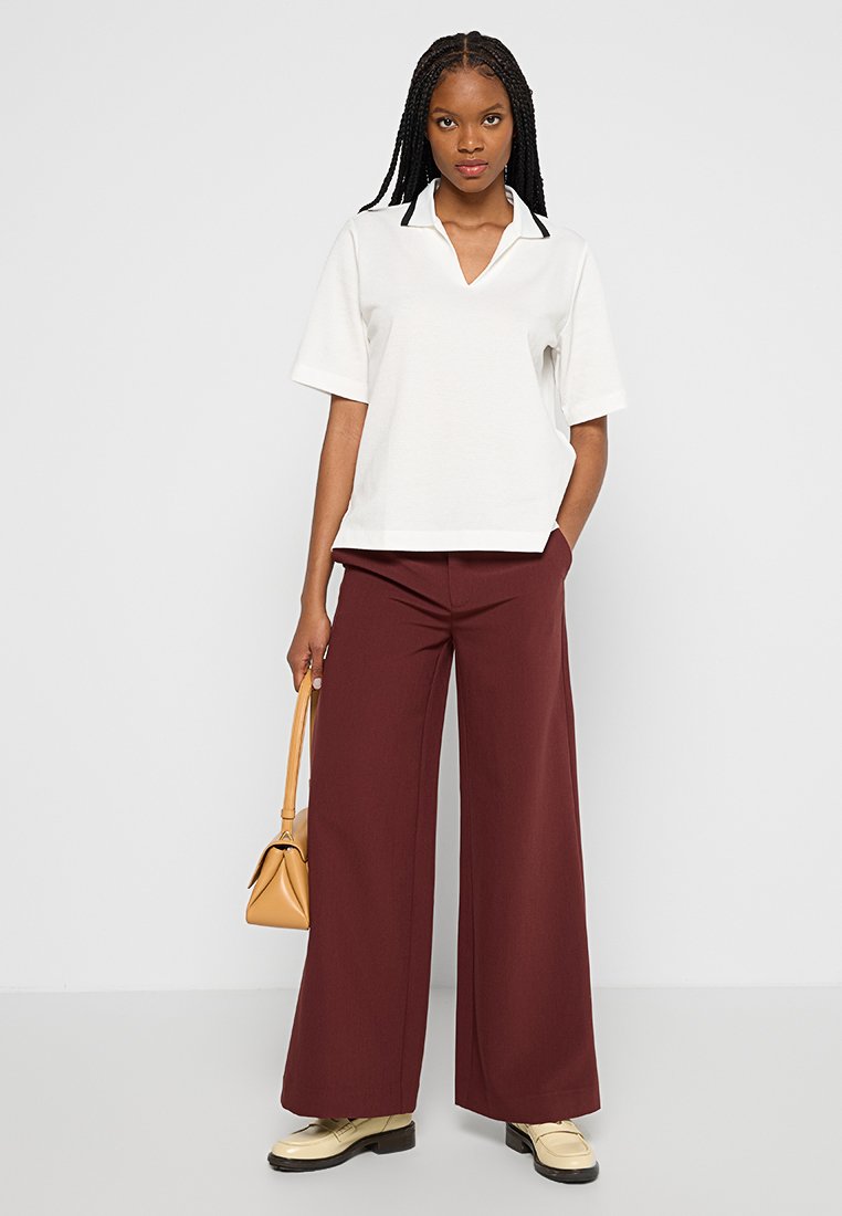 Chemise polo blanche à manches courtes et col en V, associée à un pantalon large bordeaux. Complétée par un sac à main beige et des chaussures beiges.
