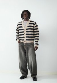 Pasiasty kardigan w kolorze kremowym i granatowym, z dekoltem w serek i guzikami, zestawiony z szerokimi szarymi dżinsami i czarnymi butami. Gładka tekstura.