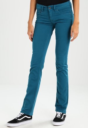 Pantalon slim fit couleur sarcelle porté par une personne avec des baskets noires et blanches, debout devant un fond clair uni.