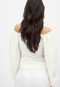 Top bianco off-the-shoulder con tessuto testurizzato, maniche a sbuffo e silhouette aderente, visto da dietro con capelli lunghi e scuri.