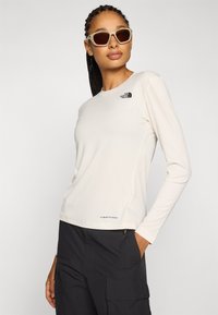 Donna con capelli intrecciati che indossa una maglia beige a maniche lunghe North Face, pantaloni cargo neri e occhiali da sole beige, in piedi con una mano in tasca.