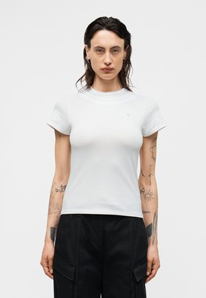 Person mit Tattoos auf den Armen trägt ein schlichtes weißes Kurzarmshirt und schwarze Hosen und steht vor einem einfarbigen hellen Hintergrund.