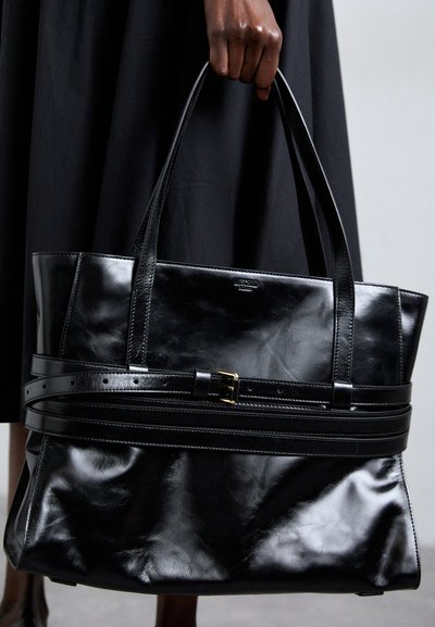 Hand holding large black leather tote bag with belt detail, worn with black skirt or dress, neutral background.