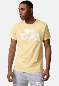 Geltonas medvilnės marškinėliai su baltu liūto grafiku ir "LONSDALE LONDON" tekstu. Trumpomis rankovėmis, apvaliu kaklu, laisvas kirpimas.