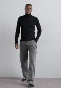 Μαύρος ζιβάγκο, γκρι χαλαρές τζιν παντελόνες, μαύρα αθλητικά παπούτσια με λευκές κορδόνες. Το μοντέλο στέκεται με άνεση με τα χέρια στις τσέπες.