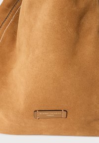 Měkká světle hnědá semišová taška s viditelným bílým prošitím a malou koženou štítkem s nápisem "Gianni Chiarini Firenze" přišitým blízko spodní části.