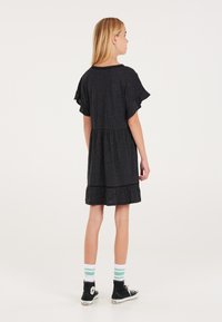 Schwarzes, kurzärmeliges Kleid mit Rüschensaum; aus weicher Stoff gefertigt. Die Accessoires umfassen gestreifte Socken und schwarze High-Top-Sneaker.
