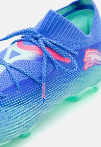 Blue football boot with a textured upper, light blue accents, pink highlights, and a snug fit collar; features a lace-up design and green sole.