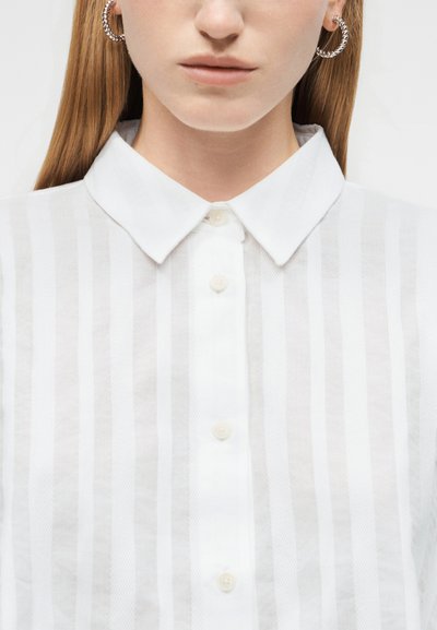 Žena v bielej pruhovanej košeli na gombíky s ostrým golierom a striebornými skrútenými náušnicami v tvare kruhu, zobrazená od krku po hornú časť hrudníka.