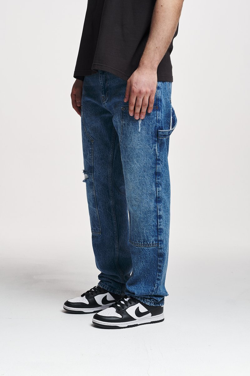 Blå denim bukser med løs passform, som har slitte detaljer og sidelommer, sammen med svarte og hvite sneakers.