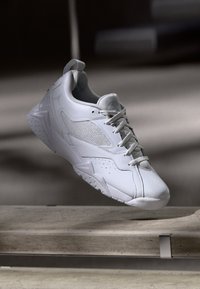 Weißer Sportschuh, der über einer Betonablage schwebt und die strukturierte Sohle sowie die Mesh-Bespanung im sanften Innenlicht präsentiert.