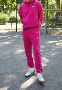 Osoba stojící venku oblečená v jasně růžové mikině Nike a teplácích s bílými a zelenými teniskami Nike na štěrkovém povrchu.