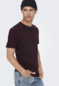 Burgundy cotton T-shirt with short sleeves, round neckline, fitted silhouette, paired with light blue denim jeans and a black beanie.