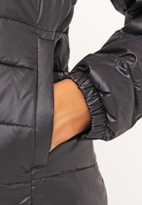 Doudoune noire matelassée avec un motif en losange, texture brillante, poignets élastiques et une poche latérale. Le tissu semble lisse et léger.