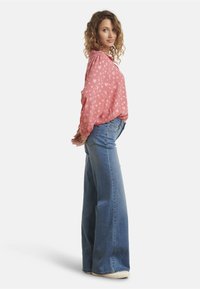 Pink blouse with white heart patterns, long sleeves, and a relaxed fit, paired with high-waisted blue flared jeans and white footwear.