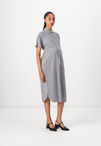 Abito a strisce bianco e nero, maniche corte, con bottoni davanti, vita con cintura, lunghezza al ginocchio. Indossato con scarpe nere con tacco.