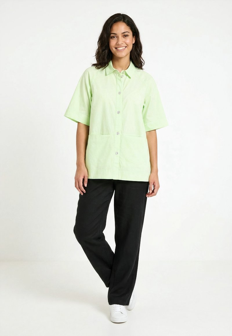 Femme debout, portant une chemise légère à manches courtes vert clair boutonnée, un pantalon noir et des baskets blanches, souriante sur un fond blanc.