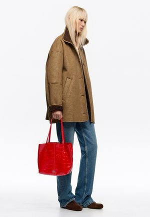 Personne portant une veste en peau retournée beige, un jean bleu et des chaussures marron, tenant un sac fourre-tout rouge brillant avec un motif texturé.