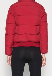 Osoba nosząca czerwoną kurtkę puchową z logo "Calvin Klein Jeans" na dole, w połączeniu z czarnymi prążkowanymi legginsami, sfotografowana od tyłu.