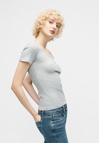 Kvinde med krøllet blondt hår iført en tætsiddende lysegrå kortærmet skjorte og blå jeans, poserer med hænderne i lommerne mod en hvid baggrund.