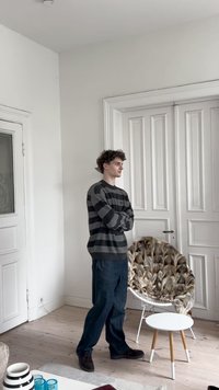 Homem de jovem com suéter riscado e calças escuras, de braços cruzados, em uma sala iluminada ao lado de uma cadeira coberta de pelagem e uma mesa pequena e branca.