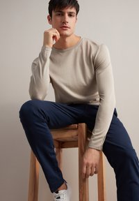 Beżowa koszula z długim rękawem, dopasowana, w połączeniu z granatowymi spodniami. Model siedzi na drewnianym stołku, w minimalistycznym tle i białych trampkach.