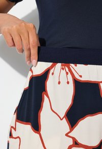 Květinová sukně s bílým základem, velkými červenými a námořnickými květinovými vzory a námořnickým pasem, doplněná námořnickou blůzou. Prst ukazuje na pas.