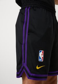 Short de sport noir avec des bandes latérales violettes et noires, logo NBA en rouge, blanc et bleu, et un swoosh Nike jaune près de l'ourlet.