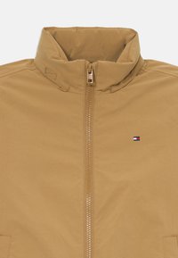 Tan zip-up jakke med høj krave og lille broderet rød, hvid og blå flaglogo på højre side af brystet.