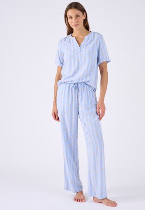 Vrouw staat blootsvoets en draagt een lichtblauw pyjamaset met verticale strepen en klein bloemmotief, kort mouwtop en losse broek.