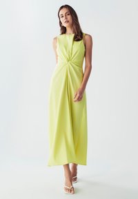 Bezginklė citrinos geltonumo suknelė, turinti priekinį mazgo akcentą, drapiruotą audinį ir laisvą siluetą, derinama su baltais sandalais su petnešėlėmis.