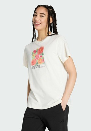 Smilende kvinde med flettet hår iført hvid T-shirt med farverigt blomsterprint og teksten "Blomster af Triumf", hænderne i lommerne på sorte bukser.