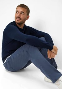 Pull en maille bleu marine, col ras du cou, manches longues, associé à un pantalon gris. Position assise avec des baskets légères. Texture du tissu lisse.