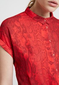 Hedvábná blouse s potiskem červeného hada, s límečkem, krátkými rukávy a uzávěrem na knoflíky. Tkanina má strukturovaný, hadí vzor pro zvýraznění detailu.