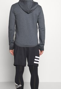 Dunkelgrauer Kapuzenpullover mit langen Ärmeln, kombiniert mit schwarzen Shorts, die an der Seite drei weiße Streifen haben, und schwarzen Kompressionsleggings.