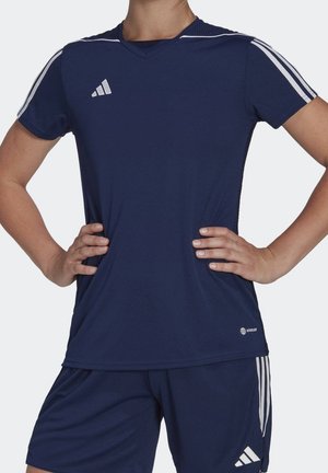 Osoba nosząca granatowy strój sportowy Adidas z białymi trzema paskami na rękawach i spodenkach, dłonie opierają się na biodrach.