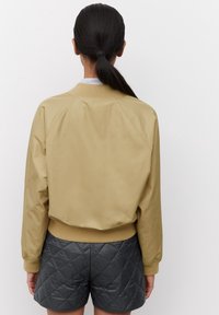 Giacca bomber beige con una texture liscia, polsini e vita a coste. Indossata sopra pantaloni corti trapuntati grigio scuro.