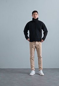 Man staat met zijn handen op de heupen, draagt een zwarte ritsopstaande trui, beige broek en witte sneakers tegen een eenvoudige grijze muur.