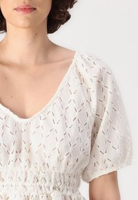 Blouse blanche en tissu texturé avec des motifs brodés complexes et une taille smockée. Les manches sont bouffantes et froncées.